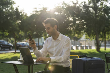 Businessman using smart phone and laptop while sitting with suitcase at sidewalk cafe - AFVF07144