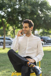 Lächelnder Geschäftsmann, der über sein Smartphone spricht, während er auf einem Stuhl vor einem Baum sitzt - AFVF07145