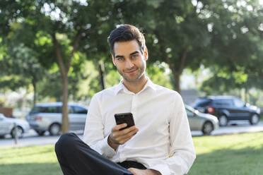 Confident businessman using smart phone while sitting against trees - AFVF07147