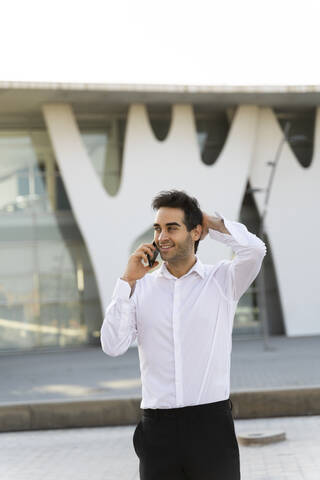 Handsome businessman with hand in hair talking over smart phone while standing against built structure stock photo