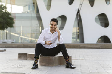 Smiling male entrepreneur with smart phone sitting on seat against built structure in city - AFVF07156