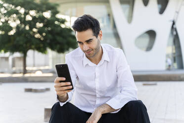 Handsome male entrepreneur using smart phone sitting against built structure in city - AFVF07158