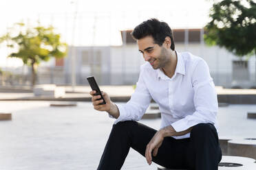 Gutaussehender Geschäftsmann, der ein Smartphone benutzt und gegen den Himmel in der Stadt sitzt - AFVF07159