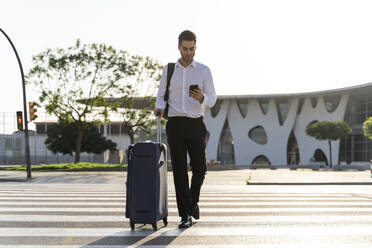 Businessman with suitcase using smart phone while crossing road against clear sky - AFVF07166