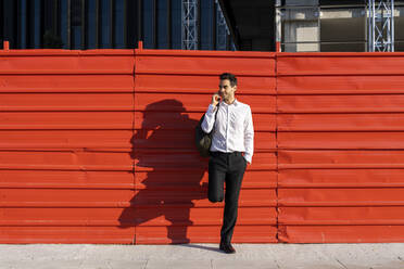 Geschäftsmann im Gespräch über Smartphone, während er an einer Wand in der Stadt an einem sonnigen Tag steht - AFVF07176
