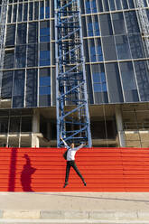 Cheerful businessman with arms raised jumping on footpath against built structure - AFVF07180
