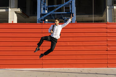 Cheerful male entrepreneur with arm raised jumping on footpath against built structure - AFVF07181