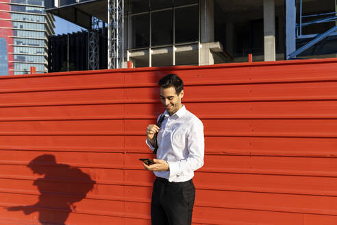 Männlicher Unternehmer, der ein Mobiltelefon benutzt, während er an einer roten Wand in der Stadt steht - AFVF07182