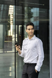 Geschäftsmann, der ein Smartphone benutzt und wegschaut, während er im Büro am Fenster steht - AFVF07190