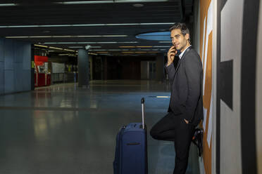Männlicher Unternehmer mit Koffer, der über sein Smartphone spricht, während er an einer Wand im Bahnhof steht - AFVF07196