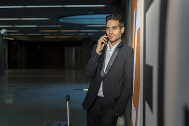Männlicher Berufstätiger mit Koffer, der über sein Smartphone spricht, während er an der Wand eines Bahnhofs steht - AFVF07197