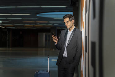 Männlicher Berufstätiger, der ein Mobiltelefon benutzt, während er mit einem Koffer an der Wand eines Bahnhofs steht - AFVF07198