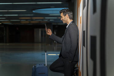 Lächelnder männlicher Unternehmer, der ein Mobiltelefon benutzt, während er mit einem Koffer an der Wand eines Bahnhofs steht - AFVF07199