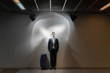 Männlicher Berufstätiger mit Maske, der mit einem Koffer an der Wand eines Bahnhofs steht - AFVF07200