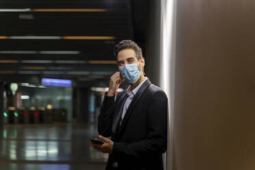 Businessman wearing mask holding smart phone while standing by wall at station - AFVF07208