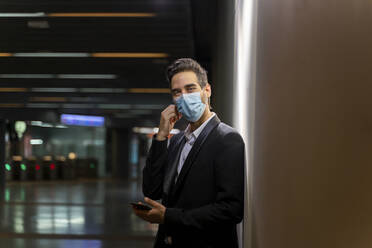 Geschäftsmann mit Maske, der sein Smartphone in der Hand hält, während er an einer Wand am Bahnhof steht - AFVF07208