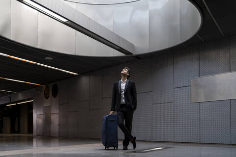Thoughtful businessman with suitcase looking through skylight while standing at station stock photo
