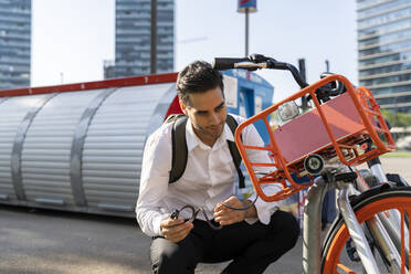 Männlicher Pendler, der in der Stadt hockend ein Elektrofahrrad repariert - AFVF07223