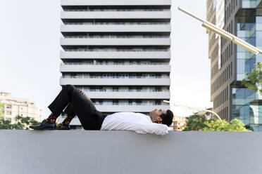 Thoughtful businessman with hands behind head lying on retaining wall in city - AFVF07227