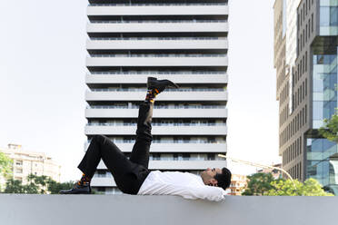 Businessman with foot up lying on retaining wall against modern building in city - AFVF07228