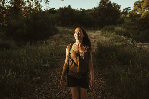Weibliche Wanderin steht im Wald, während das Sonnenlicht bei Sonnenuntergang auf ihr Gesicht fällt - DMGF00131