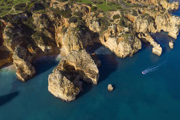 Portugal, Algarve, Lagos, Drohnenansicht eines Motorboots, das sich auf die Schornsteine der Landzunge Ponta da Piedade zubewegt - RUEF03058