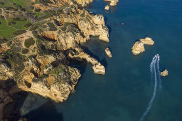 Portugal, Algarve, Lagos, Drohnenansicht eines Motorboots, das an den Schornsteinen der Landzunge Ponta da Piedade vorbeifährt - RUEF03059