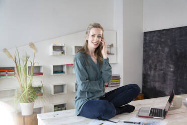 Woman sitting on desk in home office talking on the phone - FKF03796