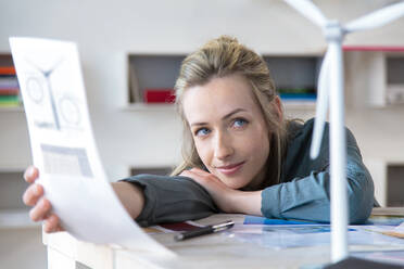 Portrait of woman leaning on desk in office with paper and wind turbine model - FKF03841