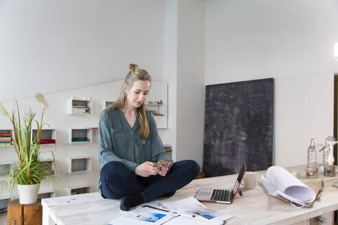 Frau sitzt auf dem Schreibtisch im Heimbüro und benutzt ihr Smartphone - FKF03848