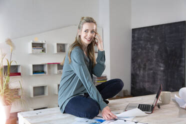 Smiling woman sitting on desk in home office talking on the phone - FKF03850