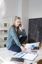 Lächelnde Frau sitzt auf dem Schreibtisch im Home Office und telefoniert - FKF03851