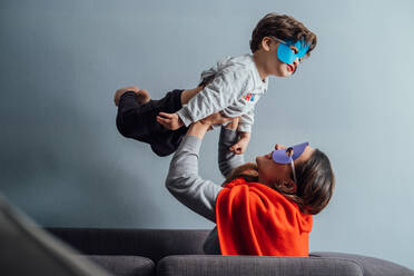 Side view of playful woman in superhero mask and cloak tossing little boy while spending time together at home and having fun - ADSF14201