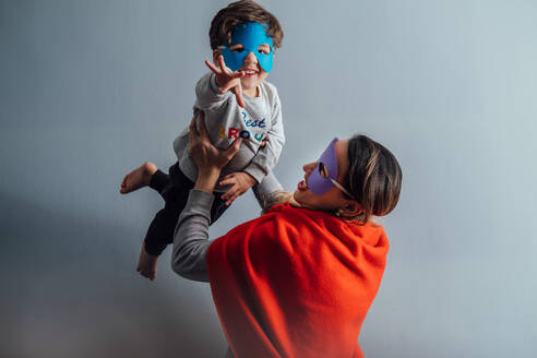 Seitenansicht einer verspielten Frau mit Superheldenmaske und Umhang, die sich mit einem kleinen Jungen vergnügt, während sie gemeinsam Zeit zu Hause verbringt - ADSF14202