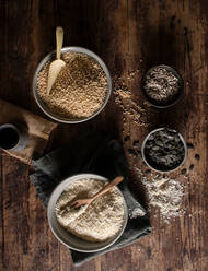 Draufsicht auf Schalen mit Hirse und Quinoa für die Zubereitung von Müsli, die auf einem Holztisch neben Servietten und Samen stehen - ADSF14269