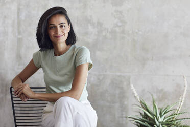 Smiling businesswoman sitting on chair against gray wall in office - MCF01195