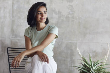 Thoughtful businesswoman smiling while sitting on chair against gray wall in office - MCF01196