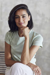 Close-up of smiling businesswoman with hand on chin sitting against wall in office - MCF01198