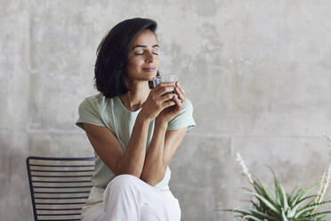 Businesswoman with eyes closed holding smoothie while sitting on chair against wall in office - MCF01202