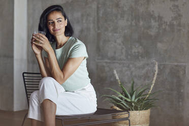 Female entrepreneur holding smoothie contemplating while sitting on chair against wall in office - MCF01205