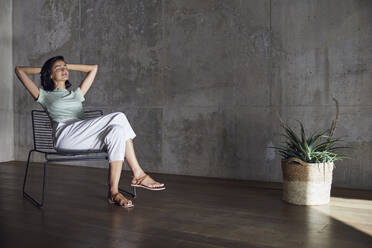 Businesswoman with hands behind head relaxing on chair against wall in office - MCF01206