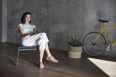 Lächelnde Geschäftsfrau entspannt sich auf einem Stuhl neben einer Pflanze und einem Fahrrad im Büro - MCF01209