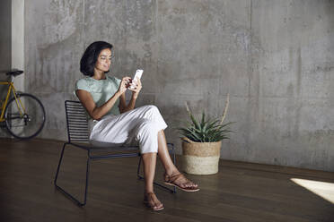 Businesswoman using smart phone while sitting on chair against wall in office - MCF01211