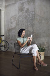 Businesswoman using mobile phone while sitting on chair against wall in office - MCF01212