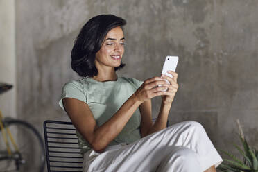 Lächelnde Unternehmerin, die ein Smartphone benutzt, während sie im Büro an der Wand sitzt - MCF01213