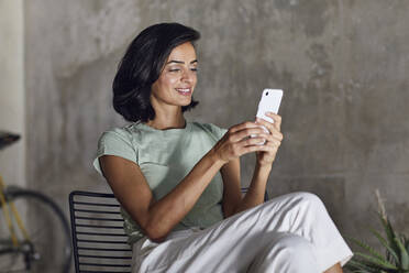 Smiling female entrepreneur using smart phone while sitting against wall in office - MCF01213