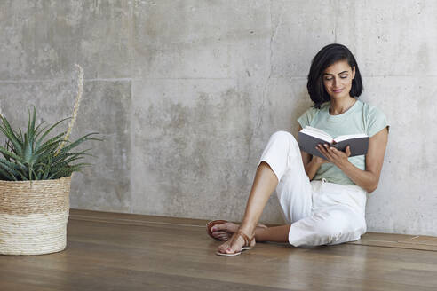 Geschäftsfrau liest ein Buch, während sie sich auf dem Hartholzboden vor der Wand im Büro ausruht - MCF01219