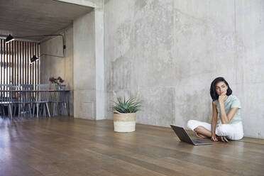 Thoughtful businesswoman sitting with laptop on hardwood floor against wall in office - MCF01226