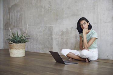 Thoughtful female entrepreneur sitting with laptop on hardwood floor against wall in office - MCF01228