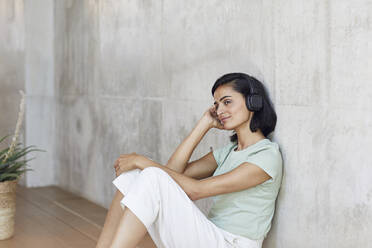 Nachdenkliche Geschäftsfrau hört Musik über Kopfhörer, während sie im Büro an der Wand sitzt - MCF01229